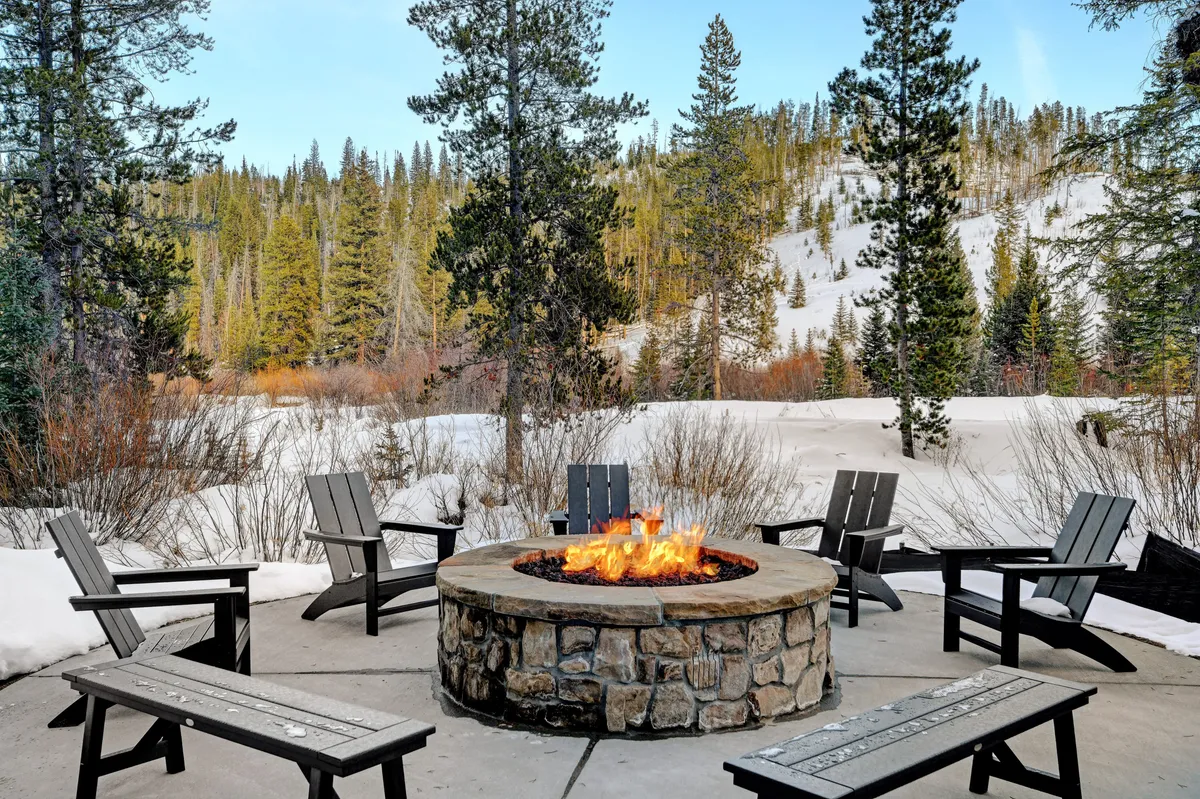 This exterior scene captures a cozy firepit surrounded by comfortable chairs, set in a snow-covered landscape framed by tall trees. The setup invites gatherings, warmth, and intimate evenings under the open sky.