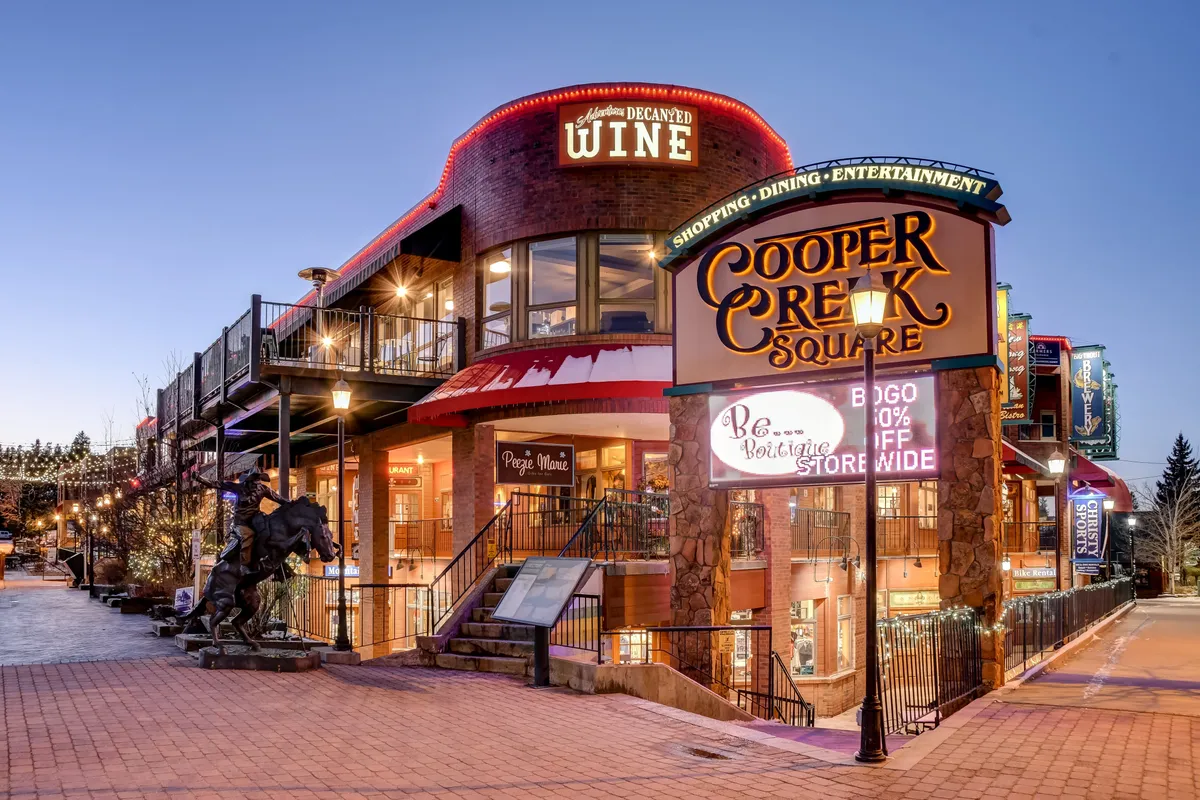 A lively shopping and dining district with the Cooper Creek Square sign, wine shop signage, outdoor seating areas, and a horse sculpture. The environment is festive, with people gathering for shopping, dining, and entertainment.