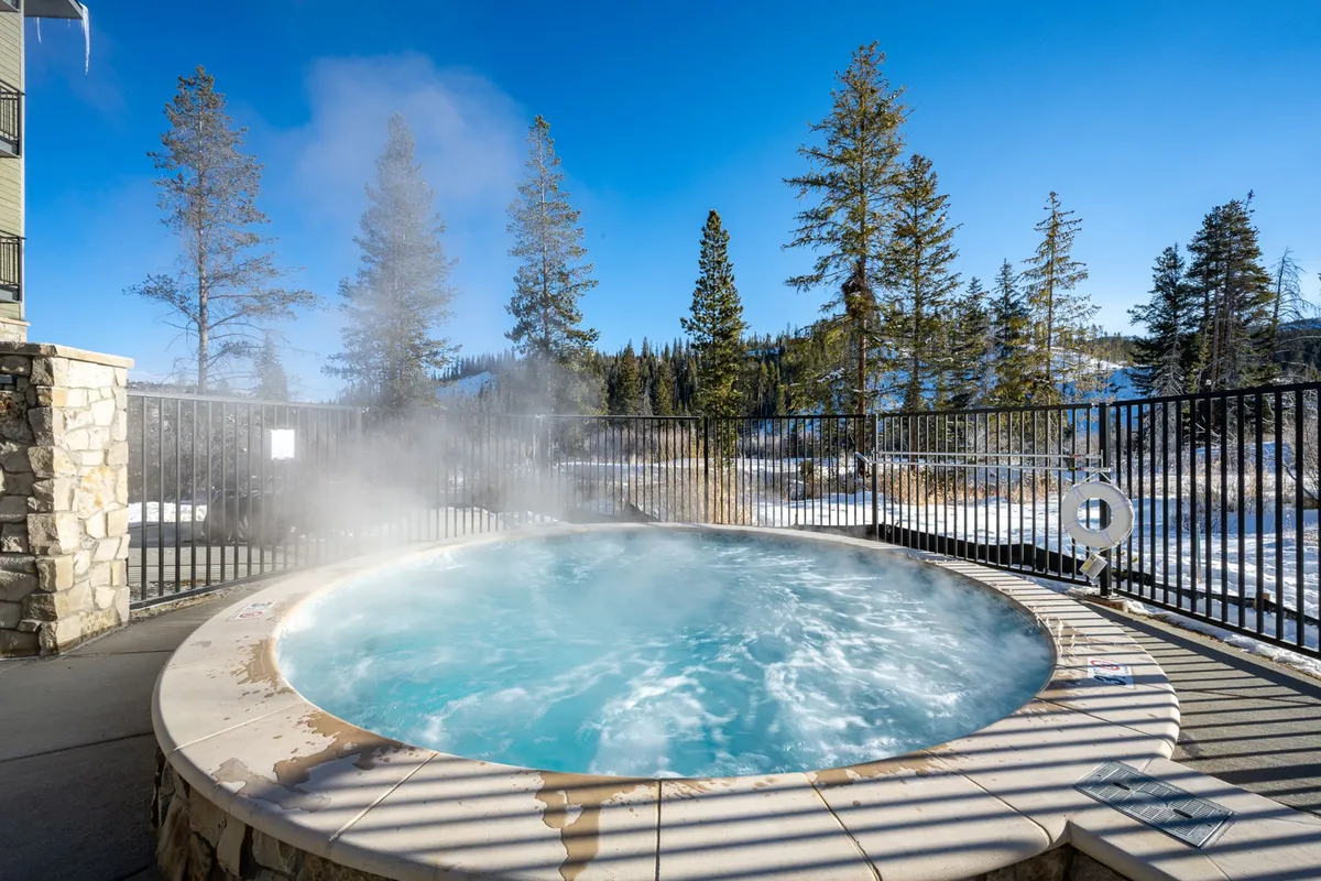 The photo highlights a steaming outdoor hot tub nestled among pine trees, with snowy mountains visible beyond. The inviting, tranquil setting emphasizes relaxation and a connection to nature, perfectly capturing the community's outdoor oasis atmosphere.
