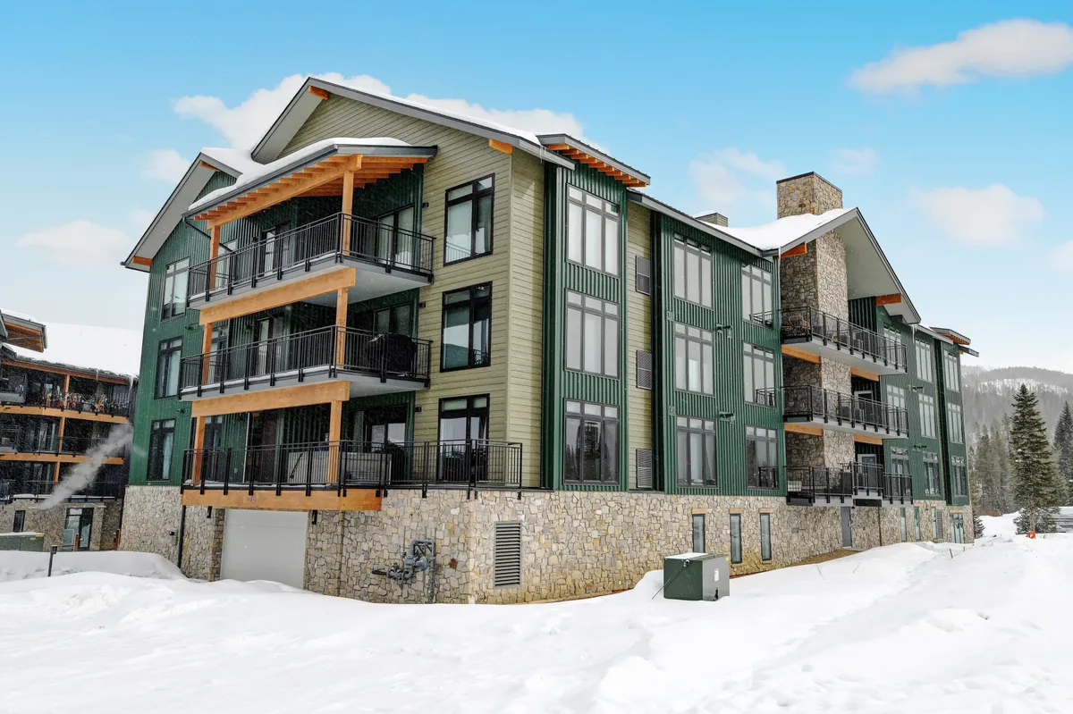 A view of the condo's exterior, featuring a stone facade, large windows, wooden balconies, and a snowy landscape. The scene is inviting and cozy, with hints of mountain adventure and relaxation.