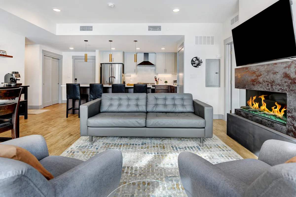 A wide-angle view encompassing the cozy living room with a plush sofa, a fireplace, kitchen bar, and dining table. The space is open and connected, filled with natural light, and offers a sense of warmth and togetherness.