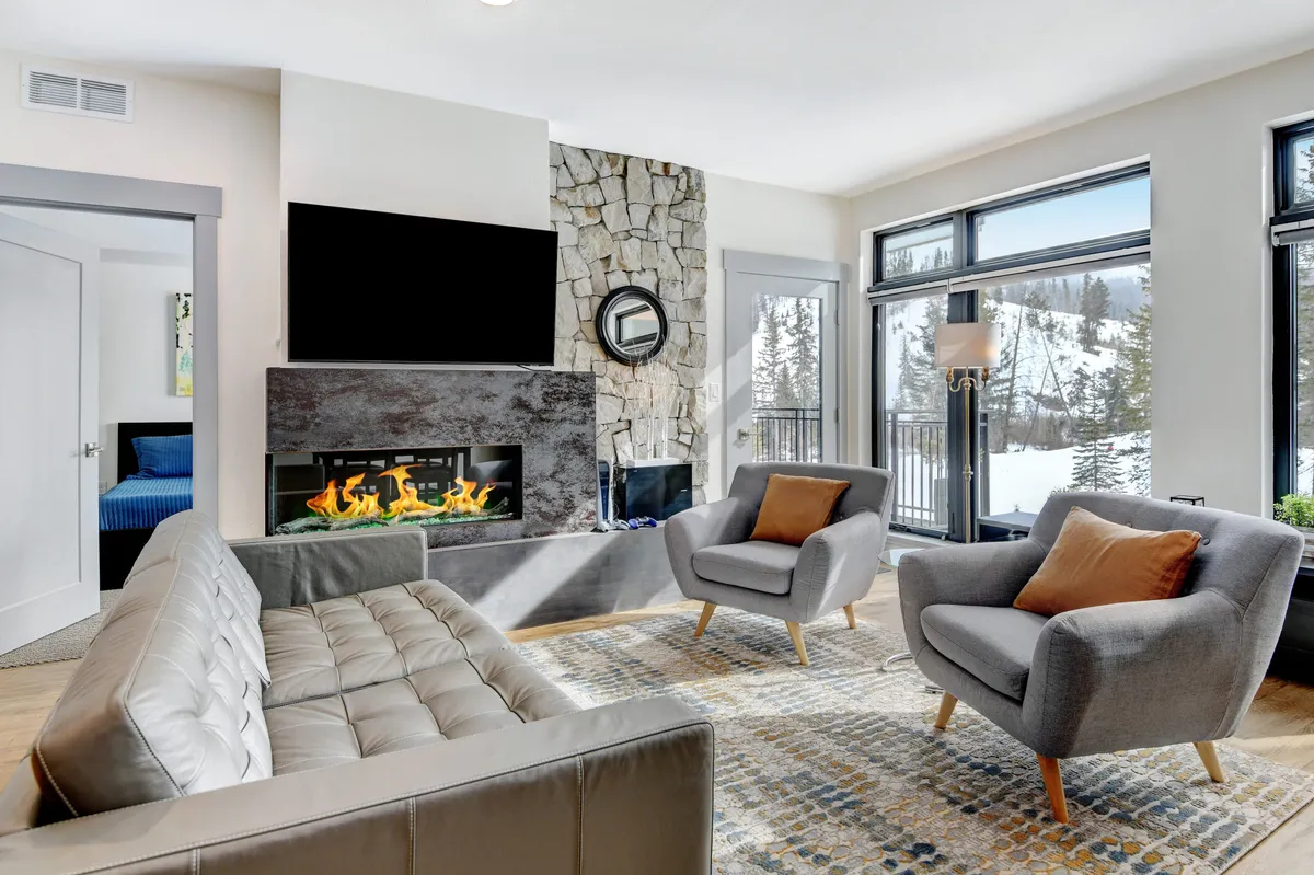 The image captures a chic, modern living area featuring a sleek fireplace at its center, flanked by plush seating and expansive windows framing scenic views of snowy Colorado nature. Soft lighting and contemporary décor create a welcoming, upscale ambiance, perfect for unwinding after a day of adventure.