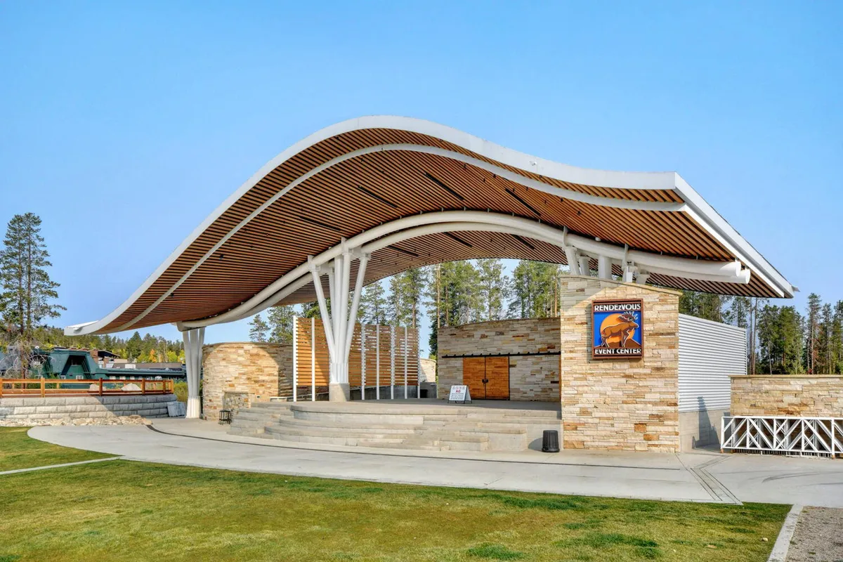 An inviting outdoor venue with a curved roof structure, a stone facade, and a designated stage area, set up for outdoor events, performances, and community gatherings. The atmosphere is festive and communal, suggesting lively happenings and shared experiences.