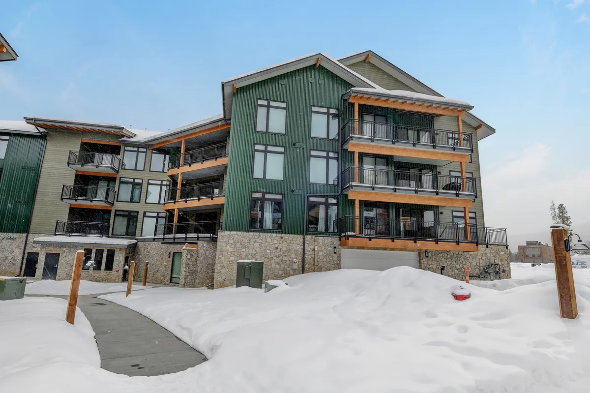 An inviting exterior shot of a green building facade with multiple balconies overlooking a snow-covered ground. People can be seen relaxing on balconies, with the surrounding winter scenery inviting thoughts of post-adventure relaxation.