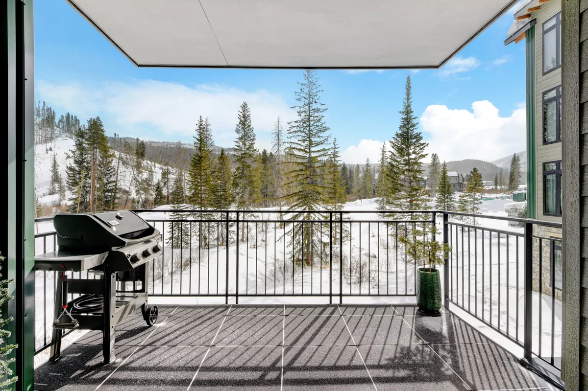 This image presents a private patio equipped with a BBQ grill, railings, and a potted plant, set against a sweeping mountain backdrop. The inviting setup hints at both relaxation and outdoor dining, with the grill and furniture suggesting social gatherings against a scenic alpine landscape.