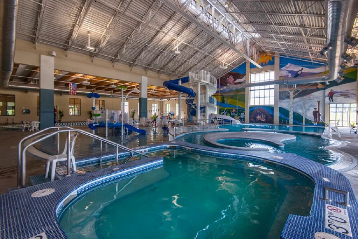 A vibrant indoor pool area at the Grand Park Rec Center, featuring water slides, a large swimming pool, colorful murals, and children playing. The joyful atmosphere and modern amenities suggest a place for both relaxation and fun.